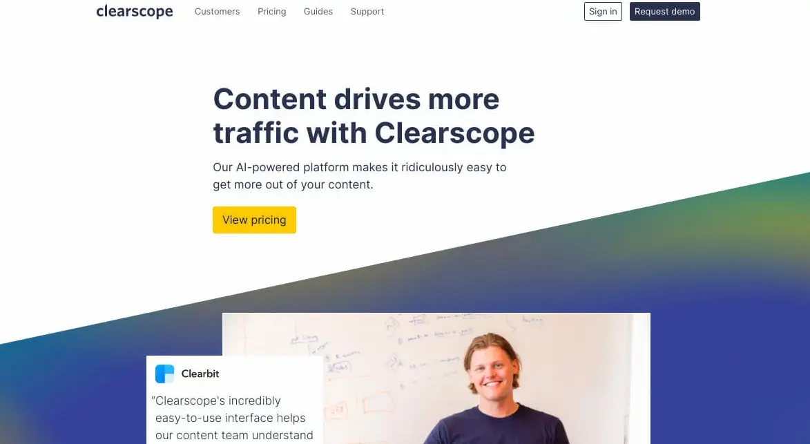 Clearscope homepage with a clean and modern design. The main headline reads 'Content drives more traffic with Clearscope.' Subheadline emphasizes that the AI-powered platform makes it easy to get more out of your content. There is a prominent yellow button labeled 'View pricing.' The top navigation menu includes options for Customers, Pricing, Guides, and Support. At the top right, there are 'Sign in' and 'Request demo' buttons. Below the main section, there is an image of a smiling person standing in front of a whiteboard, accompanied by a testimonial from Clearbit, highlighting the ease of use and helpfulness of Clearscope's interface for their content team.