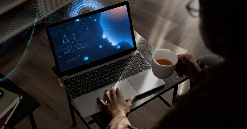 A person sitting at a table, holding a cup of coffee, and looking at a laptop screen. The screen displays a futuristic interface with the text 'A.I. Artificial Intelligence' and a stylized profile of a human head made up of digital elements. The background includes abstract digital wave patterns.