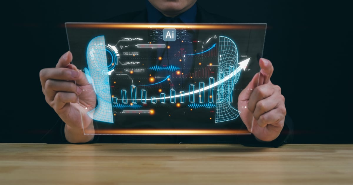 A person sitting at a table, holding a transparent digital screen displaying futuristic data and graphics related to artificial intelligence (AI). The screen shows various charts, graphs, and data points with a stylized digital head on each side and an upward trending arrow in the center. The background is dark, highlighting the illuminated elements on the screen.